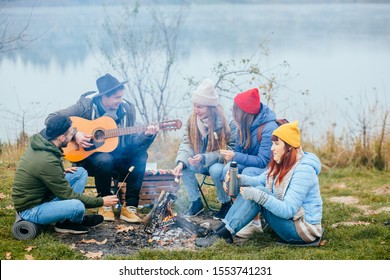 Group Friends Camping Leisure Destination Travel. Five People Sitting Around Camp Fire And Playing Guitar, Singing, Roasted Cheese, Drinking Hot Tea Tourism Relax And Travel Near The Lake In Holiday.