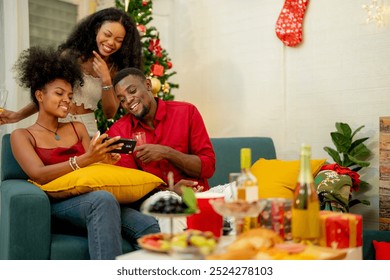 A group of friends celebrates Christmas together at home, enjoying drinks and laughter around a cozy living room. A decorated Christmas tree lights up the background, adding warmth to the holiday. - Powered by Shutterstock