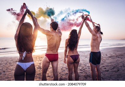 Group Of Friends Celebrating On The Beach At Sunrise. Cinematic Image Of People Having Fun On Vacation And Playing With Sparklers And Colored Smoke Bombs