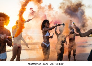 Group Of Friends Celebrating On The Beach At Sunrise. Cinematic Image Of People Having Fun On Vacation And Playing With Sparklers And Colored Smoke Bombs