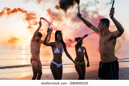 Group Of Friends Celebrating On The Beach At Sunrise. Cinematic Image Of People Having Fun On Vacation And Playing With Sparklers And Colored Smoke Bombs