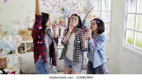 Group Of Friends Celebrating And Playing With Blower Whistles At A House Party. Young Asian Women Having Fun At A Colorful Home With Decorations And Confetti All Around. Carefree Girls Raising Hands.