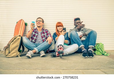 Group Of Friends Of Different Ethnics Sitting On The Street And Looking At Phone - Young Hipster People Having Fun With New Technologies - Multiracial Group Representing The Addiction To Technology