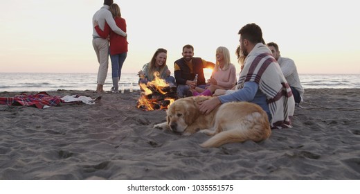 Group Of Friends With Dog Relaxing Around Bonfire On The Beach At Sunset