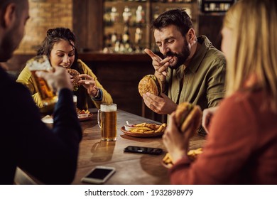 Group Of Friends Drinking Beer And Eating Hamburgers In A Pub. Focus Is On Man Tasting A Burger. 