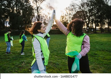 Group of friends during a volunteer garbage collection event in a park at sunset - Millennial having fun together - Happy people cleaning area with bags - Ecology concept - Two women hit high five - Powered by Shutterstock