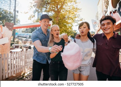 Group Of Friends Eating Cotton Candy In Amusement Park. Young Man And Women Sharing Cotton Candy Floss At Fairground.