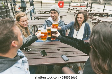 Group Of Friends Enjoying A Beer At Pub In London, Toasting And Laughing. They Are Seated Outside At A Wood Table, Wearing Winter Clothes.