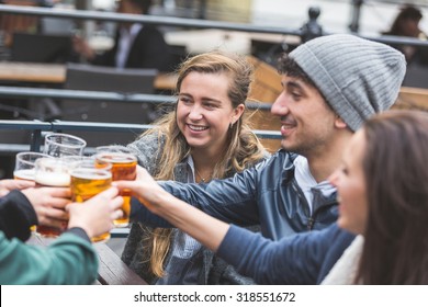 Group Of Friends Enjoying A Beer At Pub In London, Toasting And Laughing. Close Up On Two Girls And A Boy.