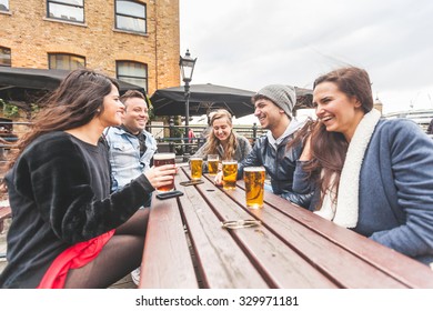 Group Of Friends Enjoying A Beer At Pub In London, Toasting And Laughing. They Are Seated Outside At A Wood Table, Wearing Winter Clothes. Friendship And Lifestyle Concepts.