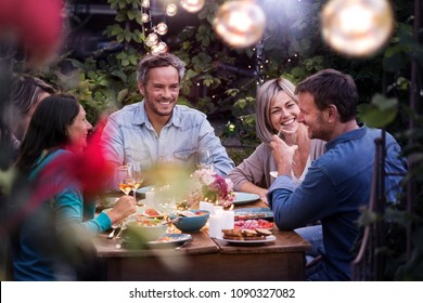 Group Of Friends Gathered Around A Table In A Garden On A Summer Evening To Share A Meal And Have A Good Time Together