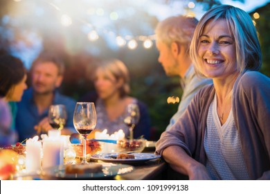 Group Of Friends Gathered Around A Table In A Garden On A Summer Evening To Share A Meal And Have A Good Time Together. Focus On A Beautiful Woman