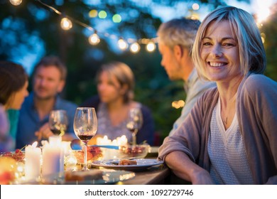 Group of friends gathered around a table in a garden on a summer evening to share a meal and have a good time together. Focus on a beautiful woman