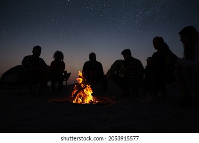 Group of friends gathering around bonfire in evening. Camping season - Shutterstock ID 2053915577