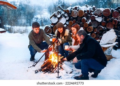 Group of friends gathering around bonfire in backyard, drinking tea and warming hands. Two happy couples relaxing and enjoying winter season while sitting around fire. Outdoor winter entertaining - Powered by Shutterstock