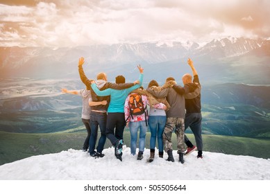 Group Of Friends Are Happy That Climbed To The Top  The Mountain. They Hug And Jump. Looking Into The Distance With Back  The Camera.