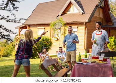 Group Of Friends Having A Backyard Barbecue Party, Grilling Meat, Drinking Beer, Playing Badminton And Having Fun On A Sunny Summer Day. Focus On The Men