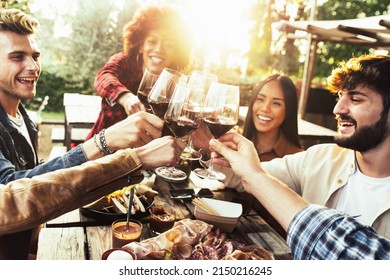 Group of friends having fun at bbq dinner in garden restaurant - Multiracial people cheering red wine sitting outside at bar table - Social gathering, youth and beverage lifestyle concept