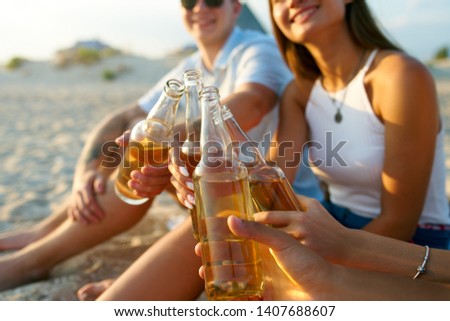 Group of friends having fun enjoying refreshing beverage and relaxing on the beach at sunset. Young men and women drink beer sitting on a sand in the warm summer evening. Isolated view of bottles.