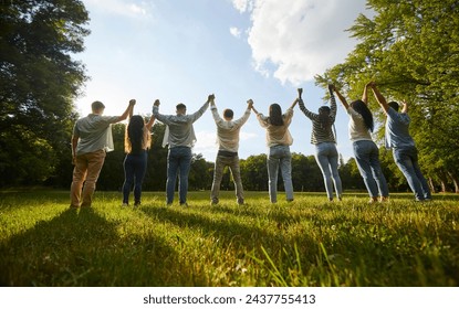 Group of friends having fun in nature. Back view team of several young diverse people standing in a row on a green grassy lawn in the park, holding hands and raising them up all together - Powered by Shutterstock