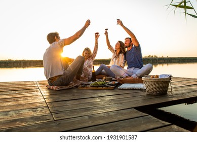 Gruppe von Freunden, die sich auf Picknick in der Nähe eines Sees amüsieren, auf Pier sitzen und Wein essen und trinken.