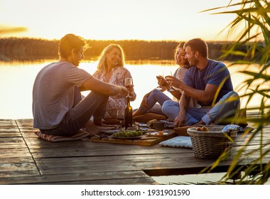 Group of friends having fun on picnic near a lake, sitting on pier eating and drinking wine. - Powered by Shutterstock