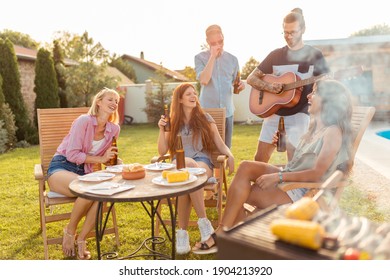 Group Of Friends Having Fun Playing The Guitar And Singing At Backyard Barbecue Party, Drinking Beer And Relaxing By The Swimming Pool