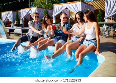 Group of friends having fun at poolside party clinking glasses with fresh cocktails splashing water by swimming pool on sunny summer day. People drinking beverages at luxury villa on tropical vacation - Powered by Shutterstock