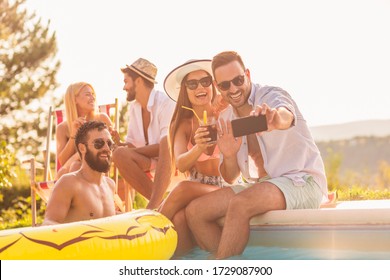 Group of friends having fun at a poolside summer party, sitting at the edge of a swimming pool, drinking cocktails and beer and taking selfies - Powered by Shutterstock