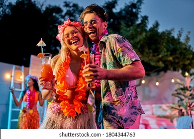 Group of friends having Hawaiian party in the evening near swimming pool. Two handsome men and three attractive young women in costumes having fun together. Summertime vibes.