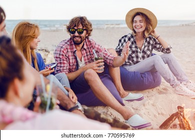 Group Of Friends Having Party Around Bonfire On The Beach At Sunset.  Hipster Man With Beard Tells Jokes. Warm Colors. Holiday Time.Sunset.