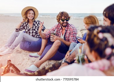 Group Of Friends Having Party Around Bonfire On The Beach At Sunset.  Hipster Man With Beard Tells Jokes. Warm Colors. Holiday Time.Sunset.