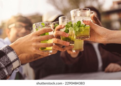 A group of friends joyfully toast with mojito glasses at an outdoor bar, with sun rays illuminating the scene and the focus on the clinking glasses. - Powered by Shutterstock