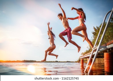 Group Of Friends Jumping Into The Lake From Wooden Pier.Having Fun On Summer Day.