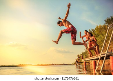 Group Of Friends Jumping Into The Lake From Wooden Pier.Having Fun On Summer Day.Sunset.