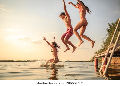 Group Of Friends Jumping Into The Lake From Wooden Pier.Having Fun On Summer Day.