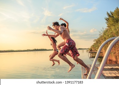 Group Of Friends Jumping Into The Lake From Wooden Pier.Having Fun On Summer Day.