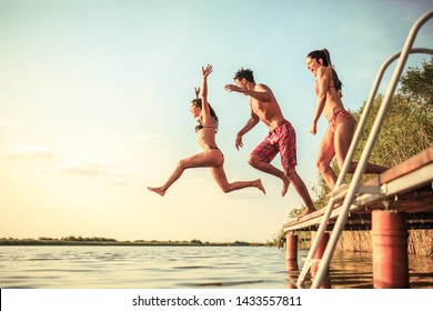 Group Of Friends Jumping Into The Lake From Wooden Pier. Summer Day Fun.