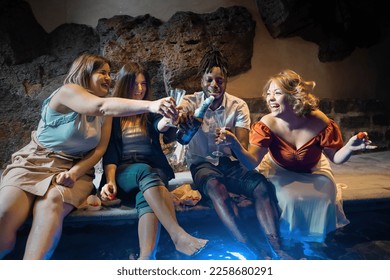 A group of friends made up of an African boy and three Caucasian women uncork a bottle of champagne at the spa pool - Powered by Shutterstock