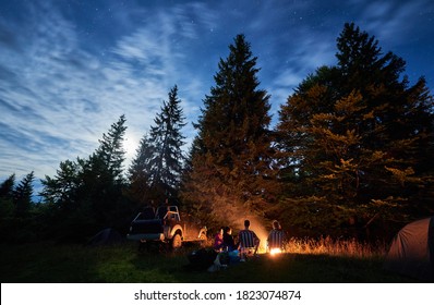 Group of friends near campfire, pickup offroad truck, tents at night camp in the mountains surrounded by spruce trees under magical starry sky, rear view. Car travel and camping concept - Powered by Shutterstock