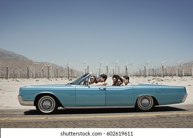 A group of friends in a pale blue convertible on the open road, driving across a dry flat plain surrounded by mountains - Powered by Shutterstock