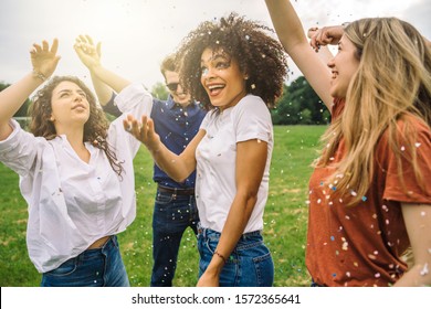 Group of friends at the park dancing under a rain of confetti - Millennials have fun in a public garden in summer at sunset listening the music - Shutterstock ID 1572365641