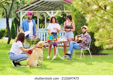 group of friends in the park at a picnic are barbecued, chatting, smiling and playing with a Labrador Retriever dog against the backdrop of greenery and a gazebo