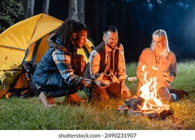 Group of friends roasting marshmallows over a campfire near a tent in the forest at night. - Powered by Shutterstock