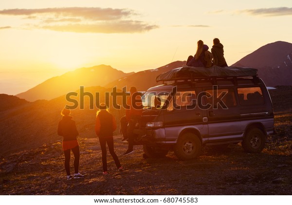 Group of friends sits on the mountain\'s top on\
background of mountains and\
sunset