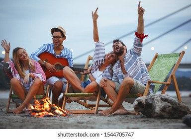 Group Of Friends Sitting Around Camp Fire At The Beach At The Autumn Evening.They Play Guitar And Singing.