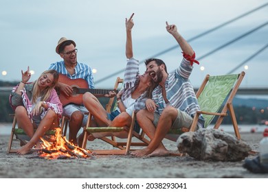 Group Of Friends Sitting Around Camp Fire At The Beach At The Autumn Evening.They Play Guitar And Singing.	
