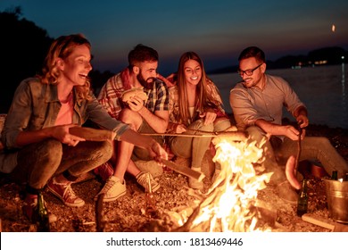 Group Of Friends Sitting Around The Fire At Night, Grilling Sausages And Having Great Time On The Beach. 