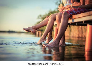 Group Of Friends Sitting And Having Fun On The Pier By The Lake On Sunset.Only Legs Are Visible.