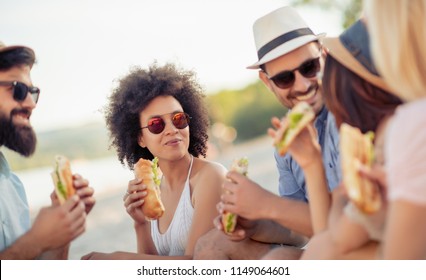 Group Of Friends Sitting On The Beach And Eating Sandwiches During Picnic.